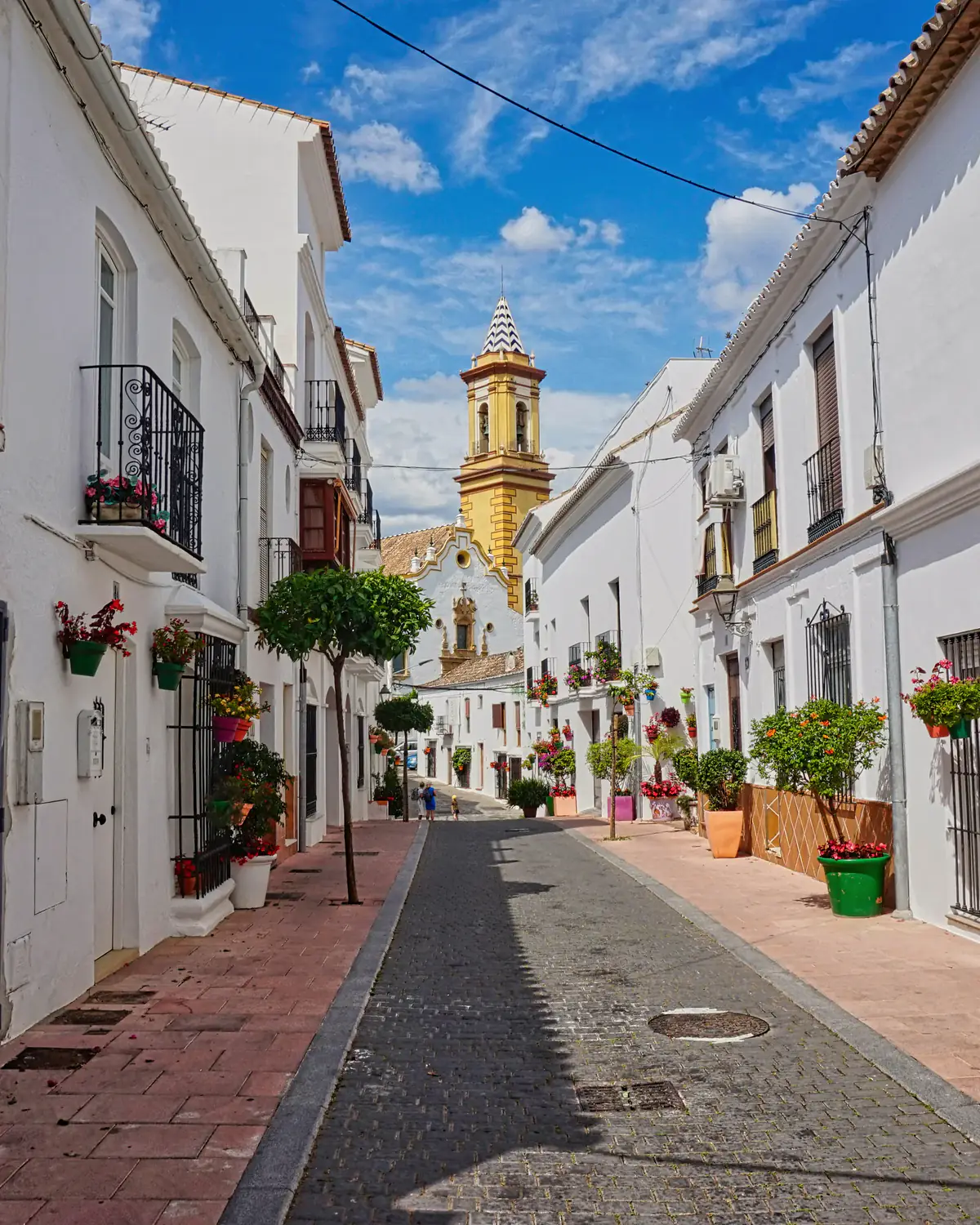 Calle encalada del casco antiguo de Estepona con macetas de flores en las fachadas y el campanario al fondo
