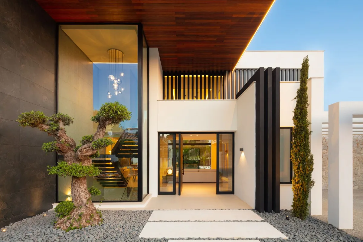 Entrada de una villa reformada en Nueva Andalucía, con un bonsái, alero de madera y puerta de cristal